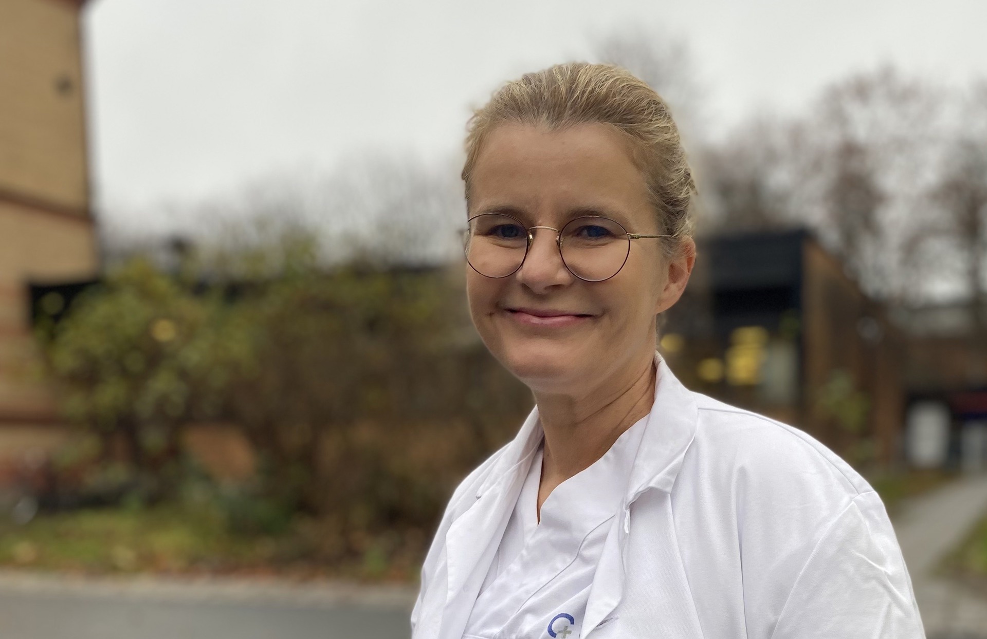 Smilende kvinne i hvitt klinikktøy. Briller. Utendørs.
