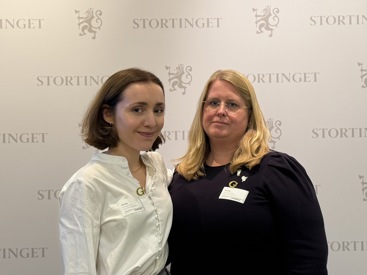 To kvinner står foran en hvit vegg som det står stortinget på, og som har stortingets logo på seg. Kvinnen til venstre er leder av Norsk medisinstudentforening, Elise Skimmeland. Hun har brunt hår til haka som er bak ørene, og hun smiler forsiktig. Hun har hvit skjorte på seg. Kvinnen til høyre er leder i Yngre legers forening, Ingeborg Henriksen. Hun har blondt hår til nedforbi skuldrene, har på seg briller og sort genser. Hun ser alvorlig ut.