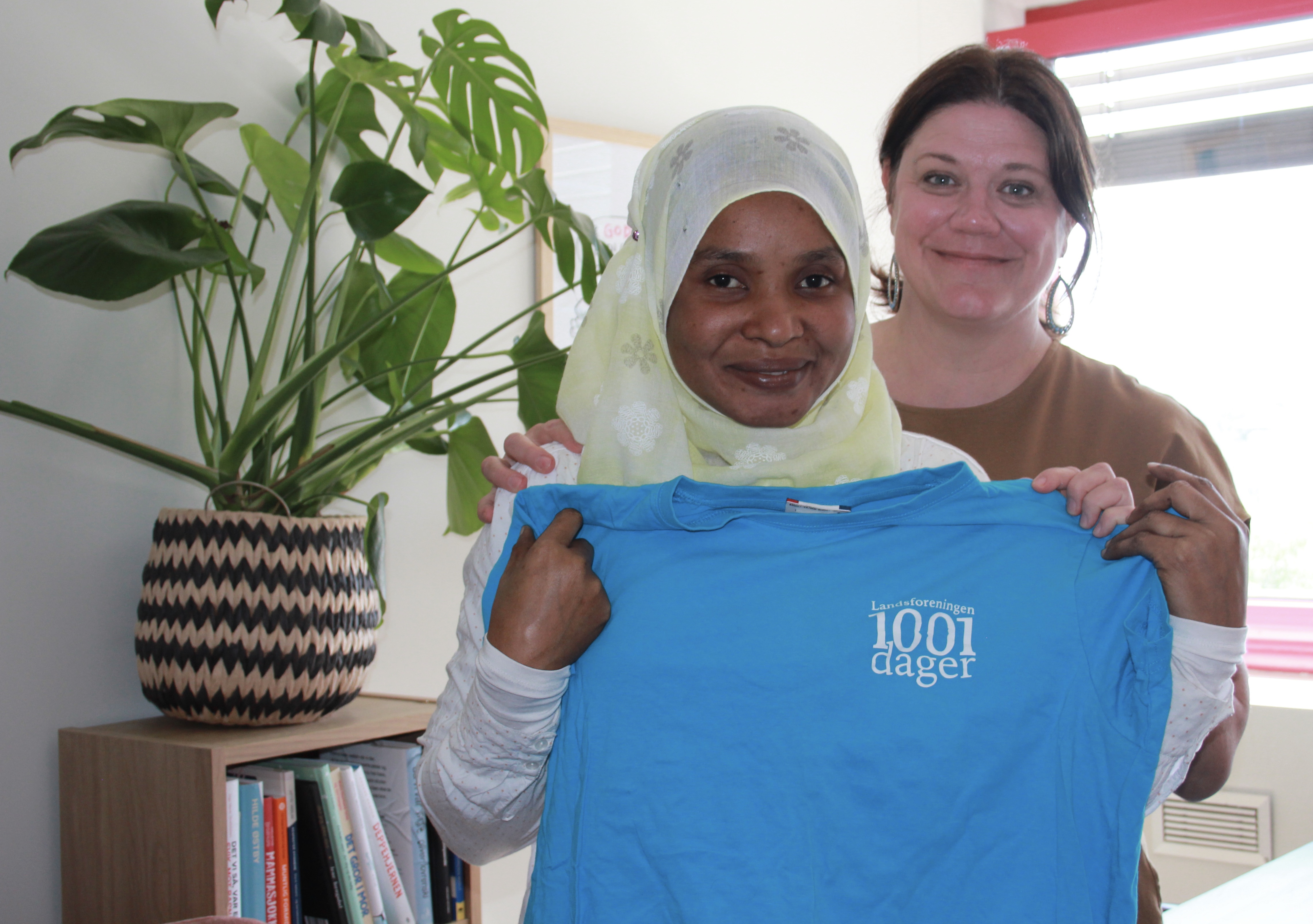En melaninrik kvinne med hodeplagg og en hvit kvinne med m&oslash;rkt h&aring;r. De holder en bl&aring; t-skjorte.
