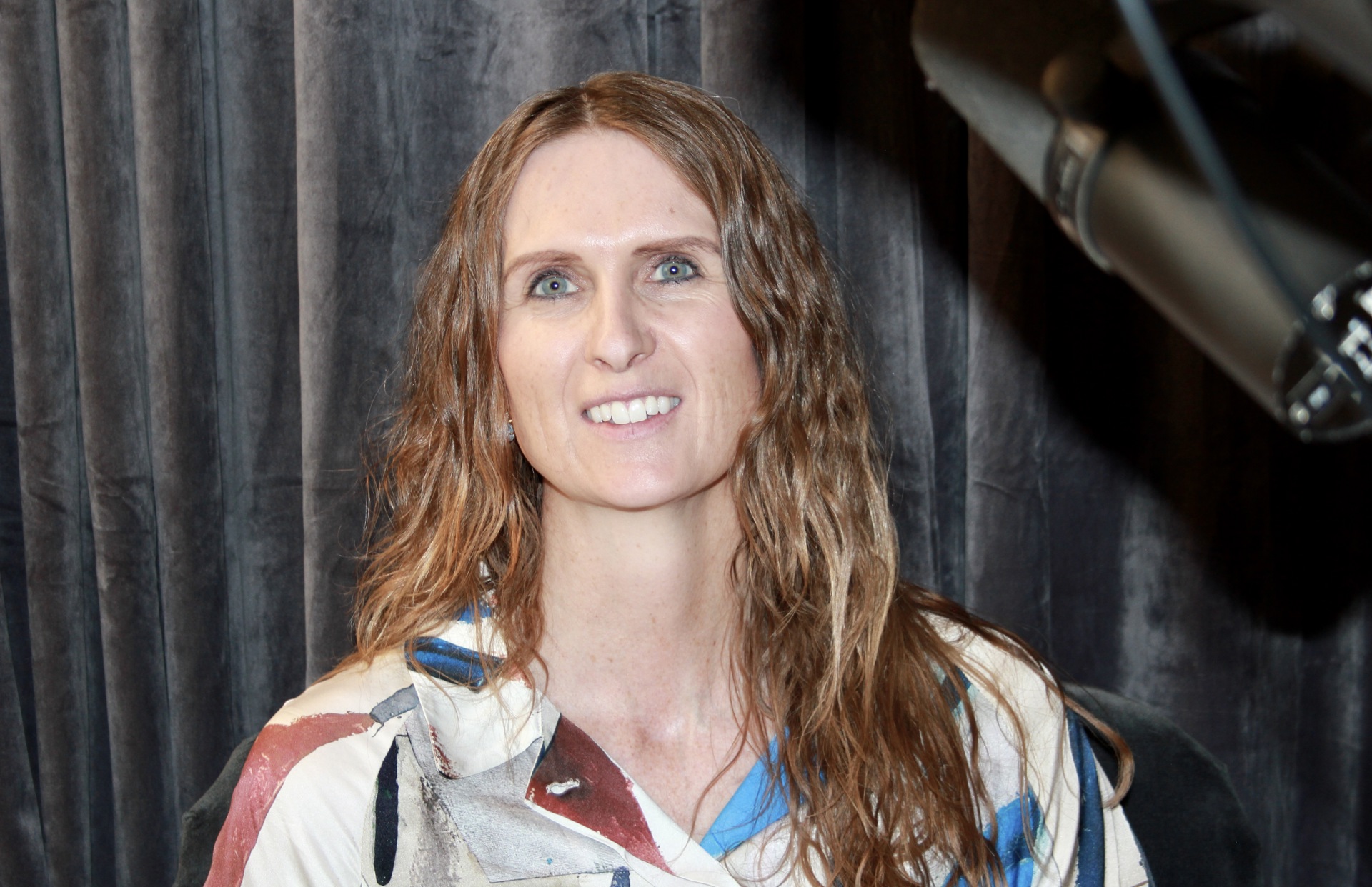 Kvinne, langt mørkeblondt hår, smiler, skjorte med blå, hvite og oransje farger. Mikrofon foran. grå fløyelsgardiner bak.