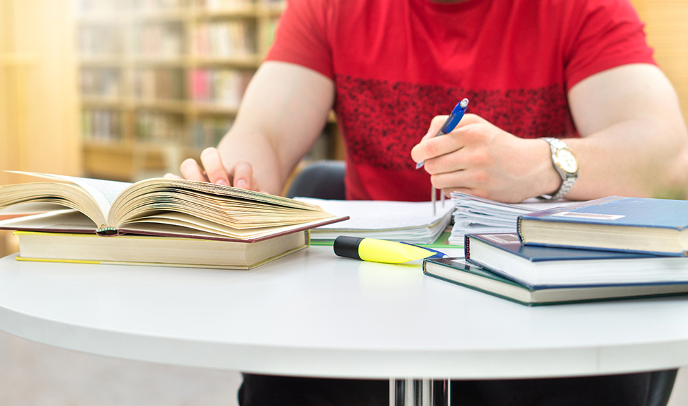  Ung student som studerer og skriver notater i offentlig eller skolebibliotek på høyskole eller universitet. Stabel og haug med bøker, penn og papir på bordet.