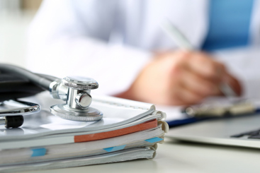 Lege som sitter ved en pult og skriver, med bøker, pc og et stetoskop i forgrunnen. Foto: Istockphoto.com