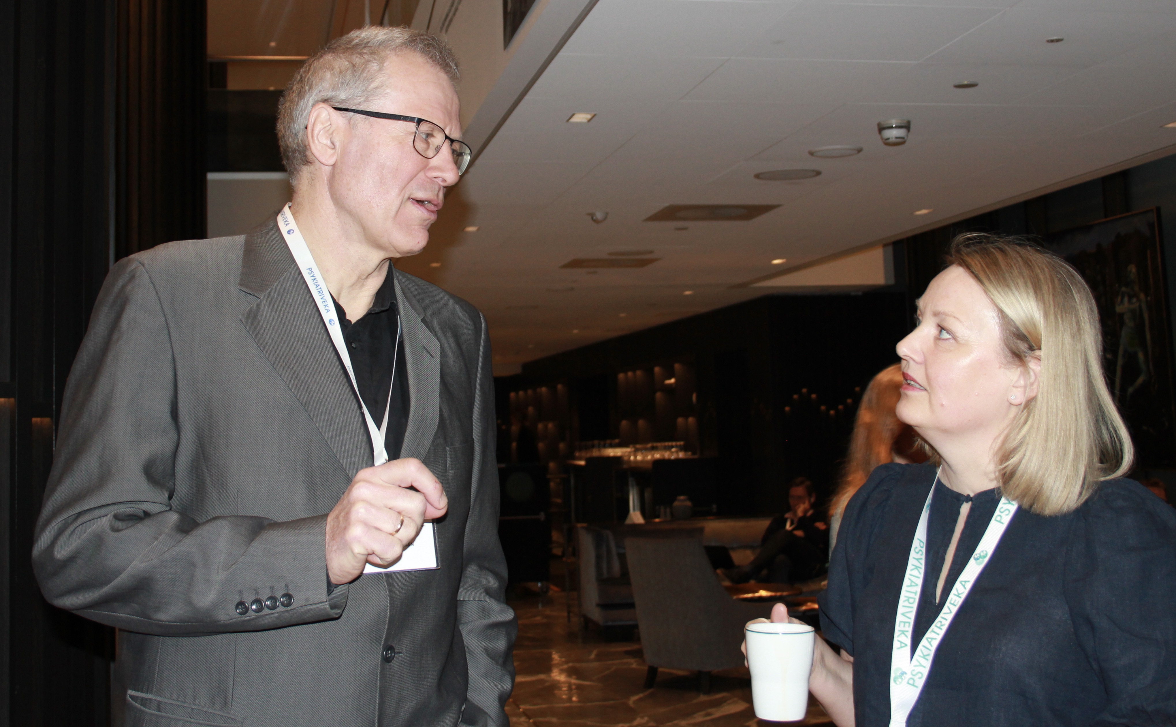 En h&oslash;y mann i beige blazer  og en kvinne med lyst h&aring;r i samtale