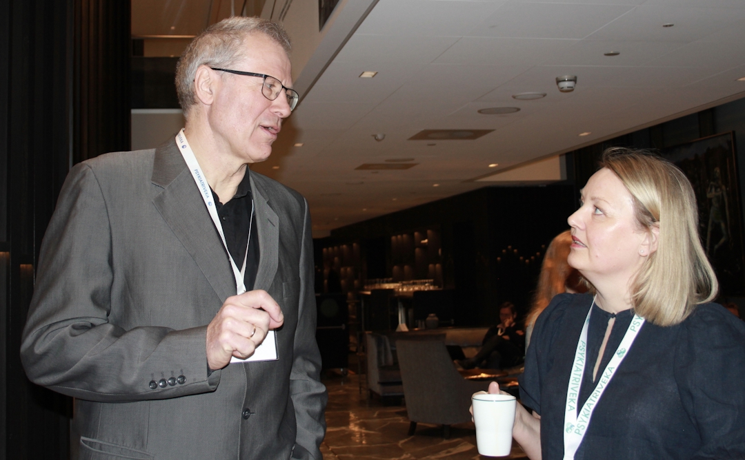 En høy mann i beige blazer og en kvinne med lyst hår i samtale