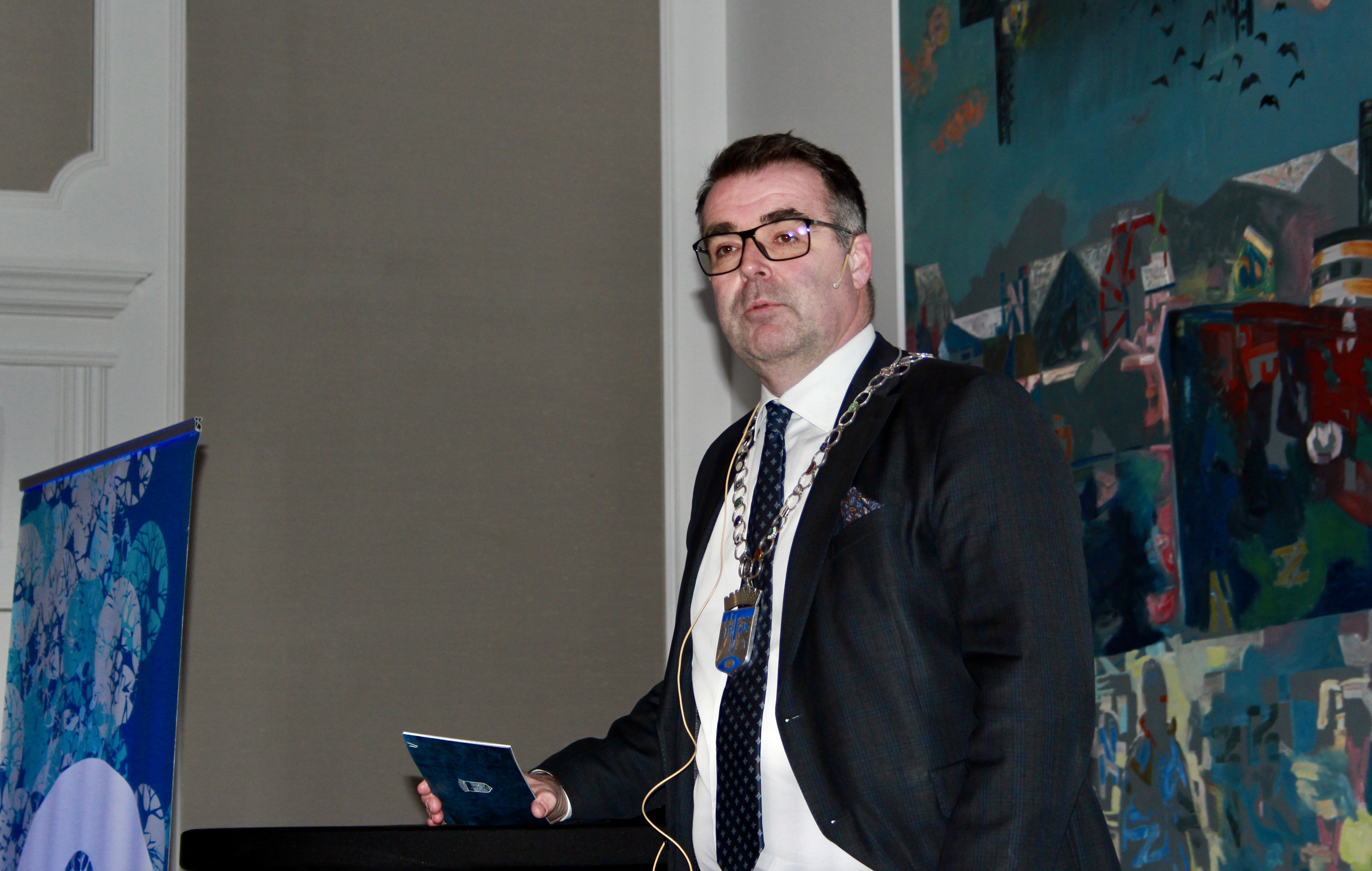 Mann med ordf&oslash;rerkjede rundt halsen, ved talerstol.