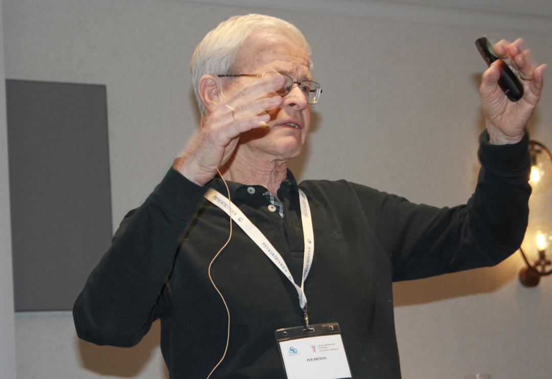 Mann som gestikulerer med armene. Mørk genser. Hvitt hår.