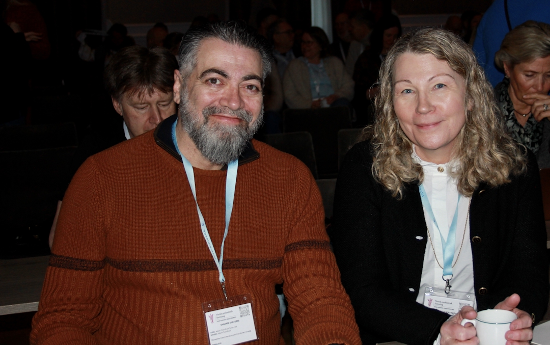 Mann mer mørkt hår og skjegg i rød genser. Kvinne med langt, lyst, krøllete hår i hvit bluse og mørk jakke