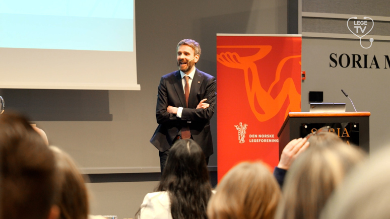 Helse- og omsorgsminister Jan Christian Vestre, står foran en forsamling. Ved siden av ham er en roll-up med logoen til Den Norske Legeforening. Han har brunt, kortklippet hår og skjegg. Han har armene i kors, og har på seg sort dress. Inni dressen er det lys skjorte med rødt slips. 