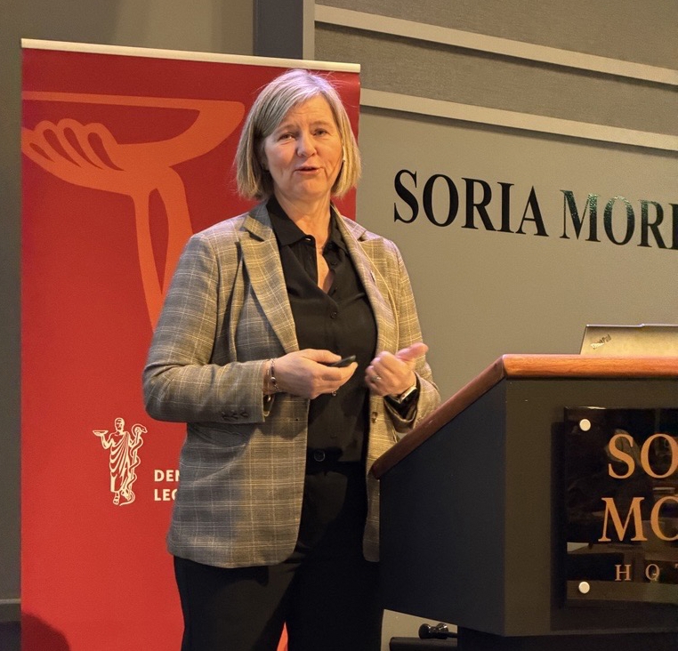 Legepresident Anne-Karin Rime står ved en talerstol. Bak henne er en rød roll-up med Legeforenignens logo. Hun har lyst hår til haken, og har på seg en brun dressjakke med ruter i. Hun har sort skjorte inni.