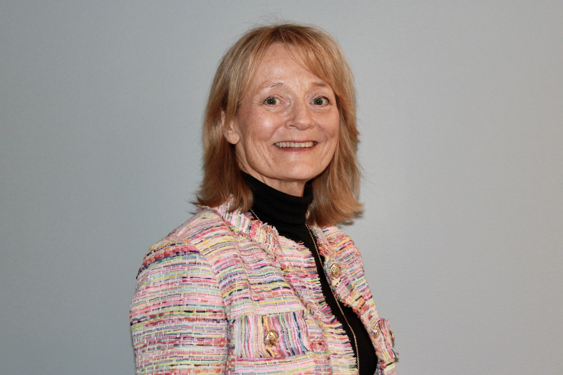 Smilende kvinne med halvlangt lyst hår. svart genser med rosa/fargerik jakke. grå bakgrunn.