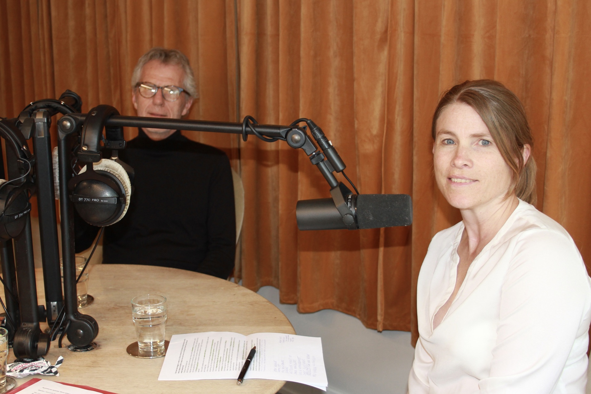 En podcaststudio med mikorfoner og høretelefoner. En mann med briller i svart genser, en kvinne med lys bluse.