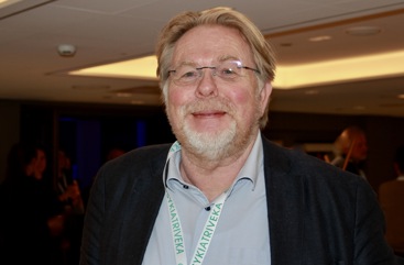 Mann som smiler, briller, skjegg med gråstenk. Lyseblå skjorte, mørk jakke.