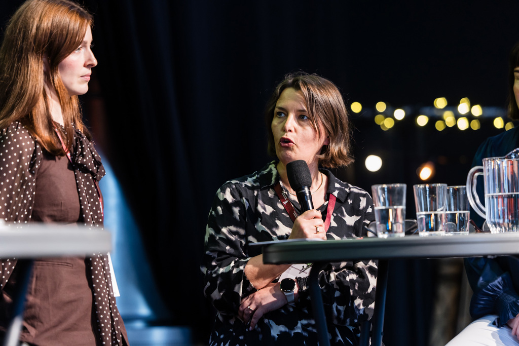 Marie Skontorp, nestleder i Overlegeforeningen, sitter ved et panelbord på en scene og holder en mikrofon mens det snakkes. Carolin Sparchholz, nestleder i Yngre legers forening, står ved siden av og vender seg mot taleren. På bordet foran dem står flere gjennomsiktige glass med vann og en vannkanne. Bakgrunnen er mørk med små punktlys som gir en scenepreget stemning.