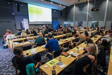 Oversiktsbilde av en konferanse med rundt hundre deltakere i en stor sal. Deltakerne sitter ved lange bord vendt mot en scene, der det holdes et innlegg foran en stor projisert presentasjon. Yngre legers forenings profilering er synlig ved scenen