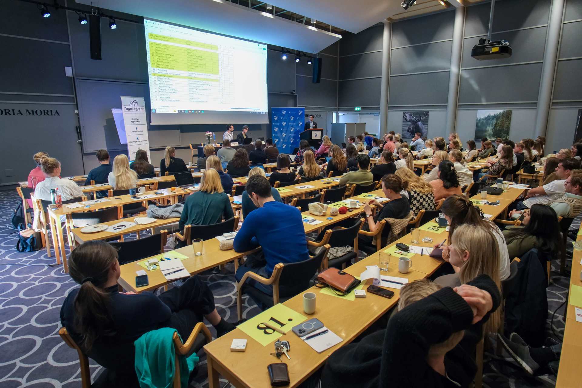 Oversiktsbilde av en konferanse med rundt hundre deltakere i en stor sal. Deltakerne sitter ved lange bord vendt mot en scene, der det holdes et innlegg foran en stor projisert presentasjon. Yngre legers forenings profilering er synlig ved scenen