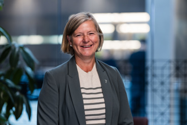 Portrettfoto av Anne-Karin Rime. Hun har kort, lyst hår, og har på seg grå blazer og lysstripet genser. Fotografert i kontormiljø med glassvegger i bakgrunnen.