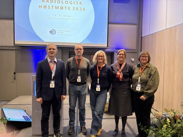 Bilde som viser fem smilende personer som står på en scene. I bakgrunnen står det "Radiologisk Høstmøte 2026" på et lærret. 