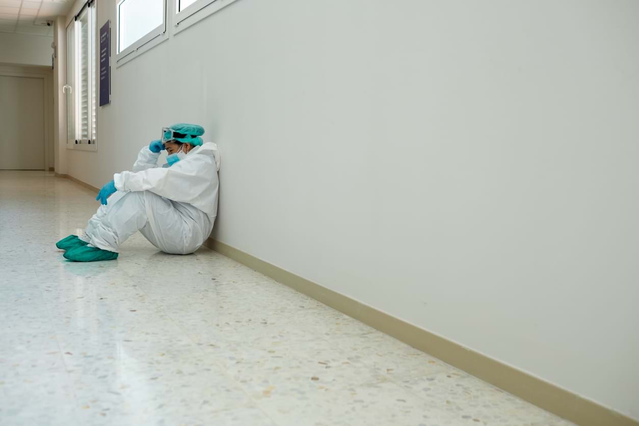 En sliten helsearbeider i beskyttelsesutstyr sittende lent opp mot en vegg i en korridor. Foto: Istockphoto.com Credit:Tempura