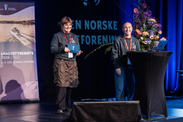 Kaja Domaas Benestad og Jeanett Myren står på en scene under et arrangement. Domaas Benestad står til venstre i bildet og holder et blått hefte med logo. Domaas Benestad har kort, rett hår som stopper rundt kjevlinjen, med en rett pannelugg. Hun har på seg en mørk genser med en liten logo på brystet, et knelangt skjørt i brun/beige tone med rynkedetaljer, mørke strømper og mørke sko. Myren står ved en svart talerstol med mikrofon og en åpen PC, omgitt av en høy blomsterdekorasjon med lilla, rosa og oransje blomster. Myren har langt hår trukket bakover og festet, slik at ansiktet er fritt. Hun har på seg en mørk genser med logo på brystet og en klassisk blå jeans. Begge har navneskilt på snor, og begge smiler. I bakgrunnen er en mørk scenebakvegg med opplyst tekst fra Den norske legeforening. Til venstre står et roll up banner med et bilde av Oslofjorden og tekst som inkluderer «Den norske legeforening» og «Landsstyremøte 2026 Oslo, 2.–4. juni, Scandic Fornebu».