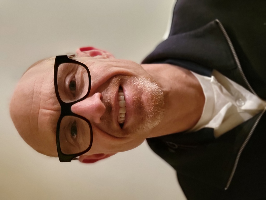 Mann med kort skjegg, bar isse. Svarte briller. Mørk genser med hvit genser inni. Smiler.
