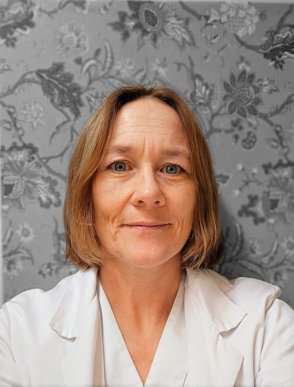 Kvinne, halvlangt blondt hår, hvitt klinikktøy. bakgrunn grå blomstrete tapet.