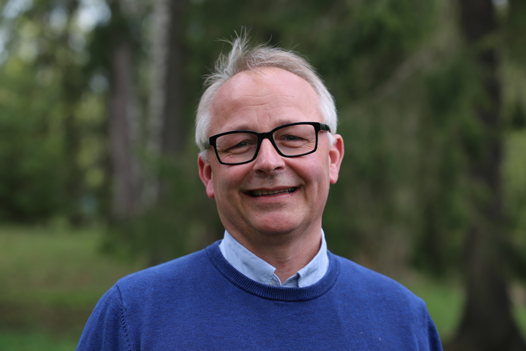 smilende mann, briller, grått hår, blå genser, lyseblå skjorte. utendørs, grønn bakgrunn