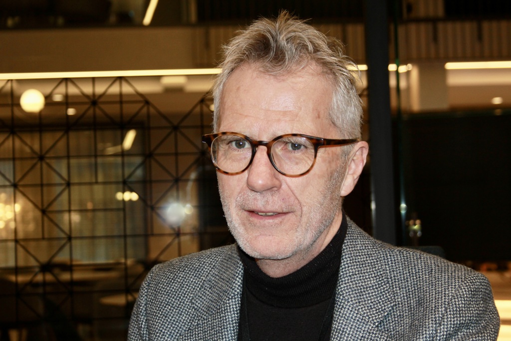 Mann, briller, grått hår, kort skjegg, dressjakke, svart genser.