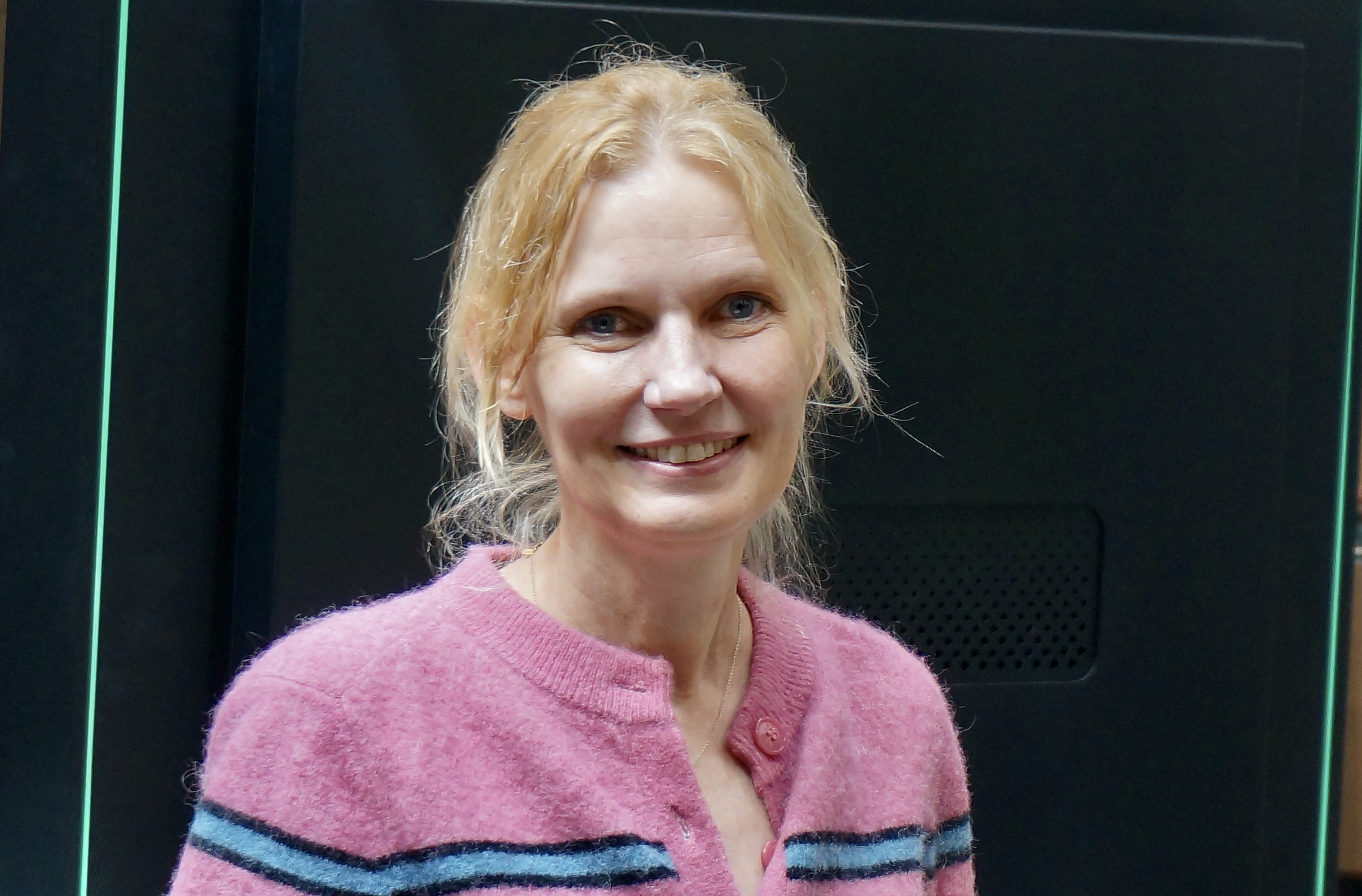 Kvinne med lyst h&aring;r. Smiler. Rosa genser. M&oslash;rk bakgrunn