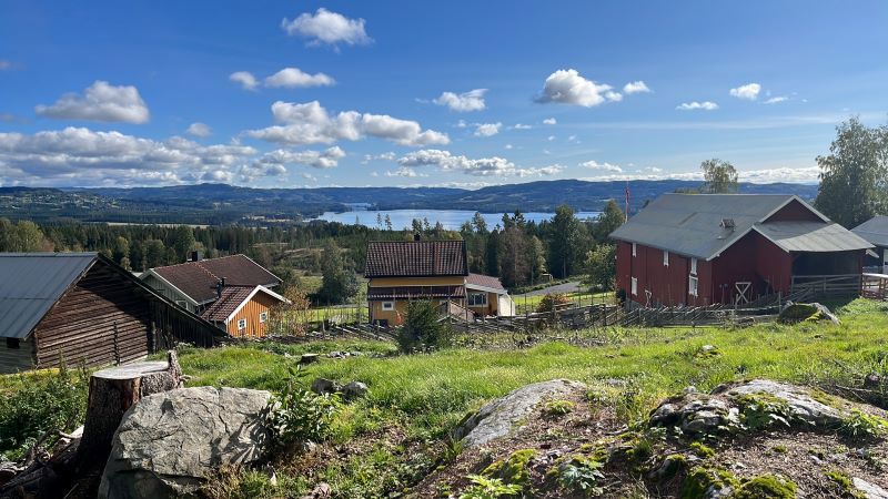 Utsiktsbilde av gårdstunet med innsjø i bakgrunnen og blå himmel med små skyer. Foto: Privat
