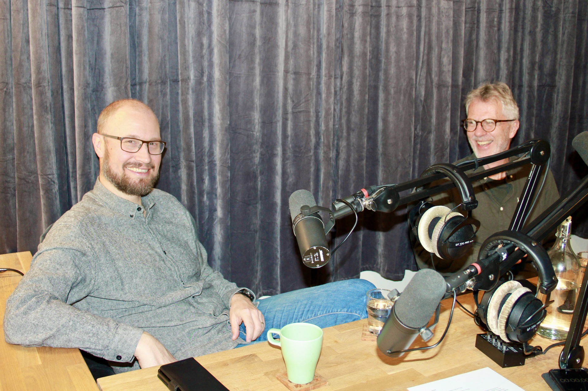 To menn med briller og skjegg bak et bord. På bordet står kaffekopper og mikrofoner.