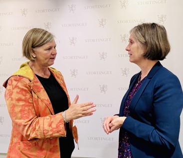 To kvinner foran en lys vegg med Stortingets logo på. F.v. President i Legeforeningen Anne-Karin Rime. Hun har lyst hår til haka, øredobber og oransje dressjakke med sort genser inni. Hun står vendt mot sidekvinnen og holder på å si noe til henne. Politiker Kjersti Toppe (Sp). Hun har også kort hår til haka, hennes er mørkt med litt grått i fronten. Hun har blå dressjakke og blomstrete skjorte inni.