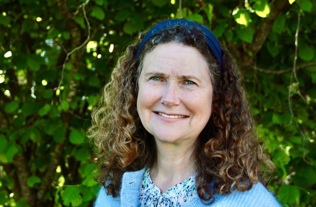 Smilende kvinne med mørkt halvlangt, krøllete hår. I bakgrunnen grønt løv.