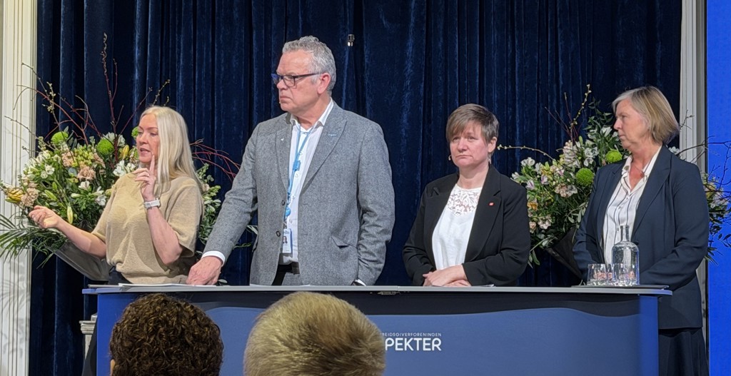 Fra venstre står forbundsleder i NSF Lill Sverresdatter Larson, leder for Delta Trond Ellefsen, forbundsleder i Fagforbundet Helene Harsvik Skeibrok og legepresident Anne-Karin Rime. Alle står foran en blå skjerm med blomster i bakgrunnen, de er bak en lang benk og vi ser dem fra livet og opp. Larsen har langt blondt hår og står halvveis vendt fra resten av panelet. Hun har beige, kortermet genser, og hun holder på å si noe. Hun har en pekefing i været, mens den andre hånden er utstrakt som klar til å ta imot den andre når hun lander poenget. Ved siden av henne står Ellefsen. Han kortklippet grått hår, svarte briller og vi ser ham halvveis i profil. Han har grå dressjakke, delta-navneskilt rundt halsen og lys skjorte. Han lene ene hånden på benken foran seg. Ved siden av ham står Skeibrok. Hun har kortklippet, brunt hår og lugg som dekker pannen. Hun ser alvorlig ut og armene er i kross oppå benken. Hun har sort jakke med Fagforbundet pin, og hvit skjorte med blondemønster i halsen. Helt til høyre står Rime. Hun har kort, lyst hår til haka, og ser mot Larsen som snakker. Vi ser henne i profil. Hun har blå dressjakke og skjørt, og en hvit skjorte med mørkeblå striper. Foran hendene hennes som hviler på benken, står en vannflaske og noen glass.   