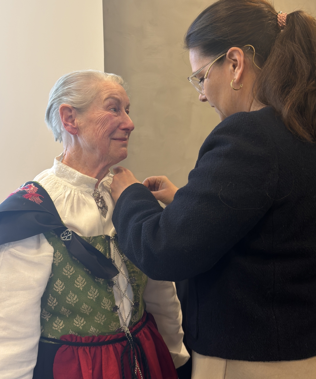 Kirkengen, en eldre dame med kortklippet hår, står i tradisjonell bunad vendt mot en kvinne med langt, brunt hår i hestehale (representant fra slottet) som fester en medalje eller utmerkelse ved kragen. Bildet er tatt innendørs mot en lys vegg, og viser en nær situasjon der hendene arbeider med festet på plagget.