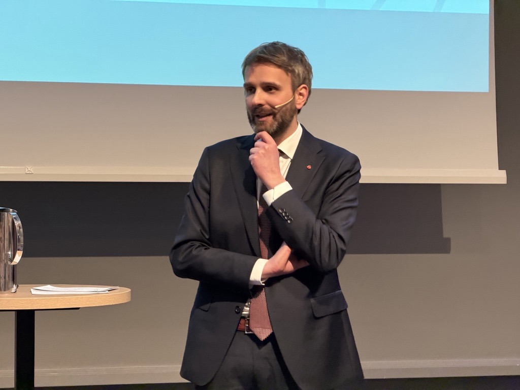 Helse- og omsorgsminister Jan Christian Vestre står ved siden av et lite bord. Han har kortklippet, brunt hår og skjegg. Han har på seg mørk dress, og står med en arm over magen, en hånd til haken. Inni dressen har han hvit skjorte og rødt slips.