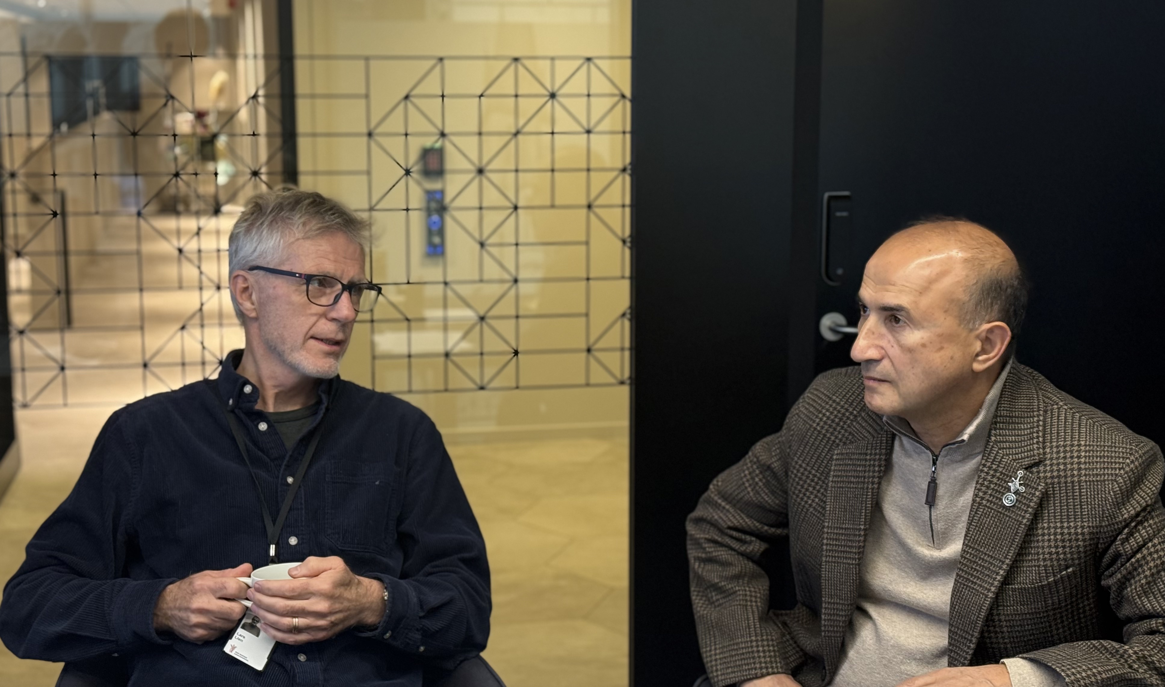 To menn sitter ved et bord. En i m&oslash;rk skjorte, en i lys genser.