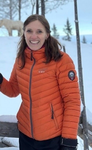 Bilde av Torhild Hovdal i orange dunjakke utendørs med en hest i bakgrunnen. Foto: Privat