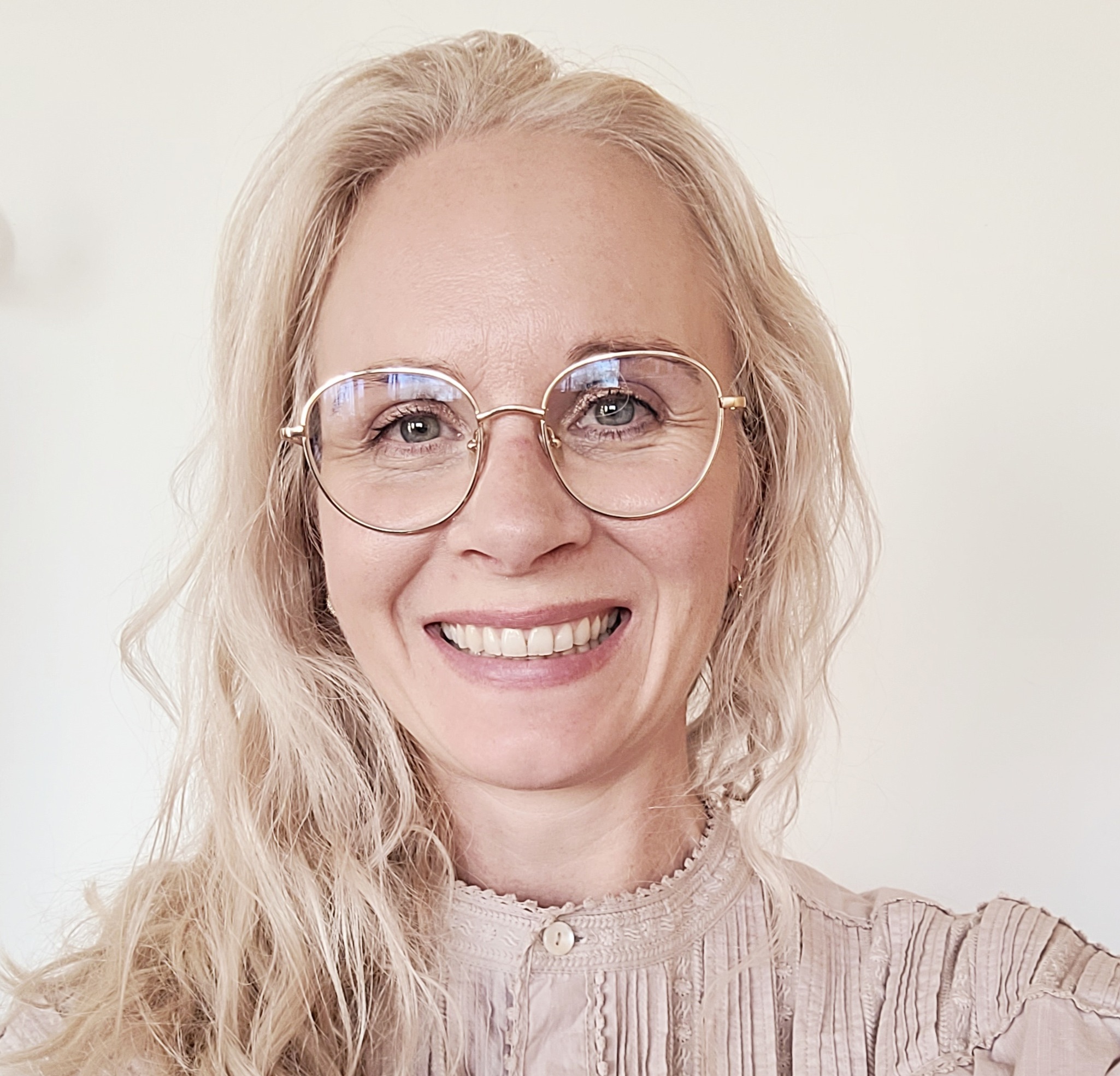 Smilende kvinne med briller og lyst, langt h&aring;r. Lys topp.