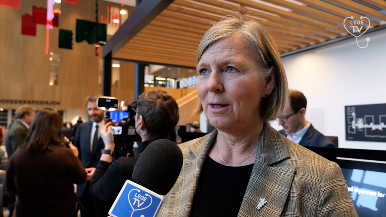 Legepresident Anne-Karin Rime. Hun blir intervjuet, og foran henne er en mikrofon med "Lege-TV"-logo på. Rime har lyst hår til haka som hun har bak ørene. Hun har på seg en brun dressjakke med lyse firkanter i stoffet. Hun har sort topp inni, og på høyre jakkeslag er Legeforeningens pin.