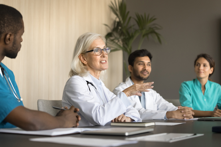 Kvinnelig lege som snakker til helsepersonell rundt et bord. Foto: Istockphoto.com/fizkes